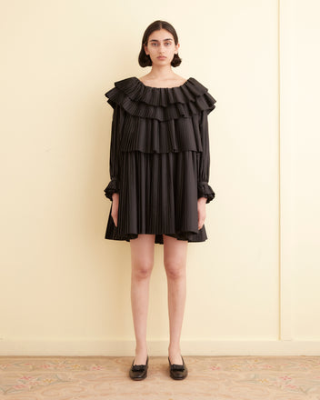 A woman in a Brunch Dress - Black, showcasing cascading pleats and ruffles, stands in a room with light-colored walls. Made from recycled polyester, this elegant outfit embodies both sophistication and sustainability.