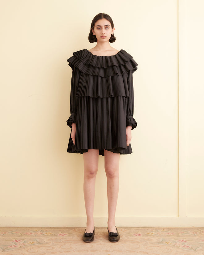 A woman in a Brunch Dress - Black, showcasing cascading pleats and ruffles, stands in a room with light-colored walls. Made from recycled polyester, this elegant outfit embodies both sophistication and sustainability.