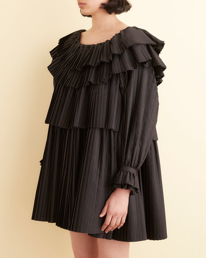 A woman in a Brunch Dress - Black, showcasing cascading pleats and ruffles, stands in a room with light-colored walls. Made from recycled polyester, this elegant outfit embodies both sophistication and sustainability.