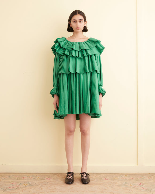 A person stands against a beige wall wearing the Brunch Dress - Green, which is made from 100% recycled polyester and features cascading tiers of pleats, paired with black shoes.