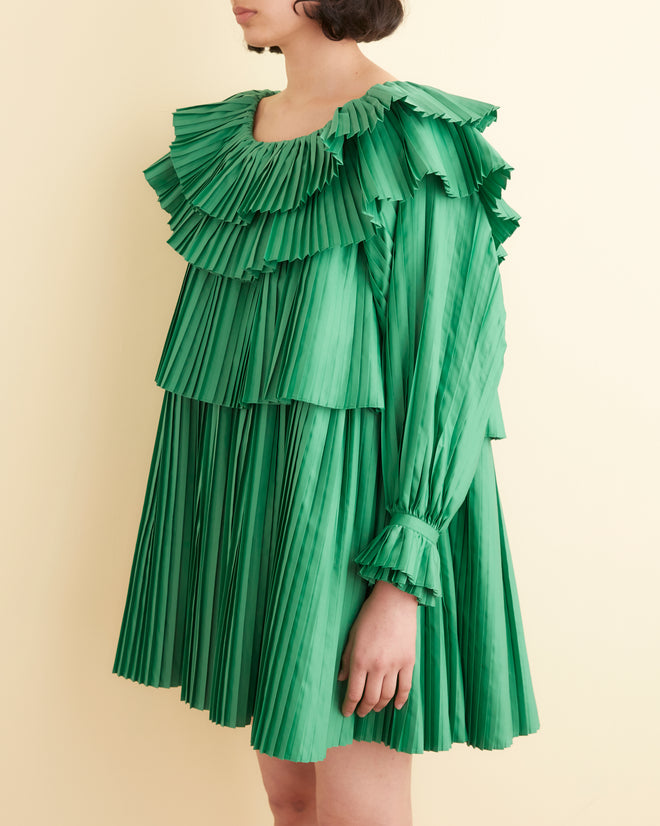 A person stands against a beige wall wearing the Brunch Dress - Green, which is made from 100% recycled polyester and features cascading tiers of pleats, paired with black shoes.