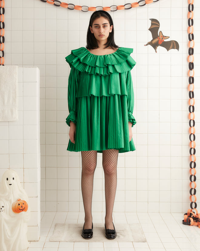 A person stands against a beige wall wearing the Brunch Dress - Green, which is made from 100% recycled polyester and features cascading tiers of pleats, paired with black shoes.