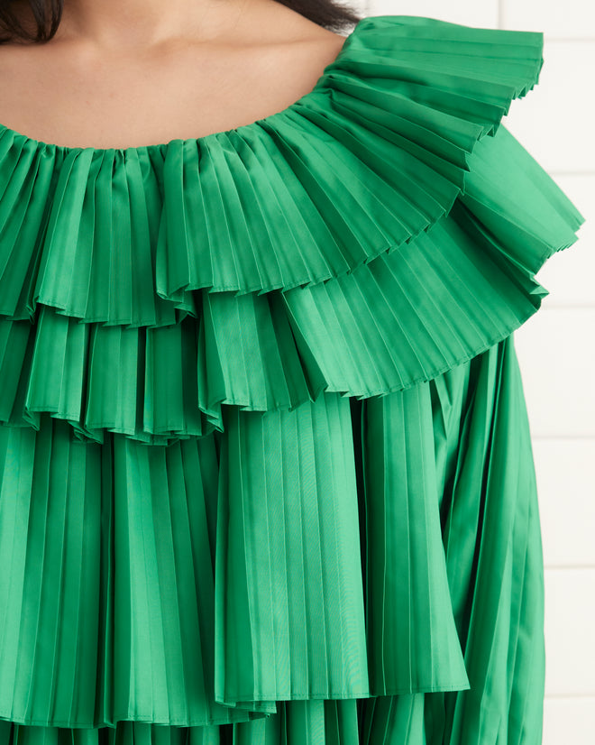 A person stands against a beige wall wearing the Brunch Dress - Green, which is made from 100% recycled polyester and features cascading tiers of pleats, paired with black shoes.