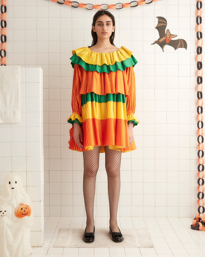 A person is standing in a room, wearing the Brunch Dress - Multi, a knee-length 1970s inspired dress featuring orange, yellow, and green ruffled tiers with long sleeves. They are also donning black shoes.