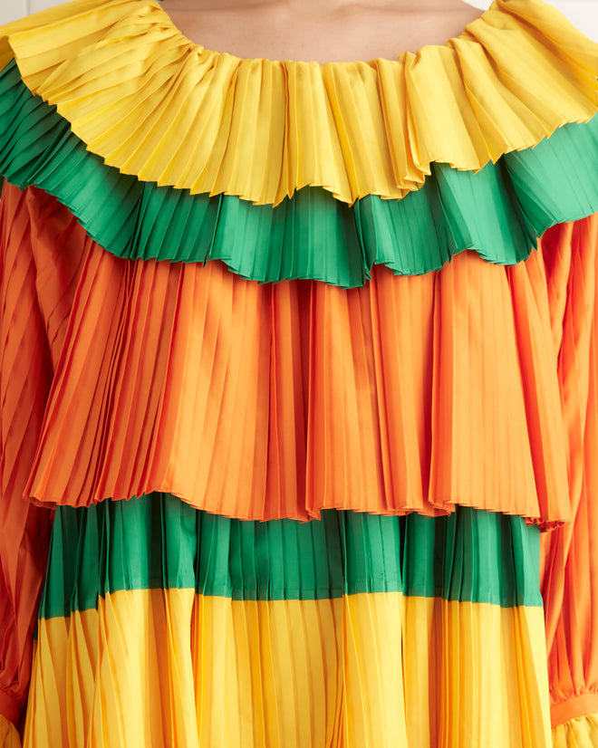 A person is standing in a room, wearing the Brunch Dress - Multi, a knee-length 1970s inspired dress featuring orange, yellow, and green ruffled tiers with long sleeves. They are also donning black shoes.