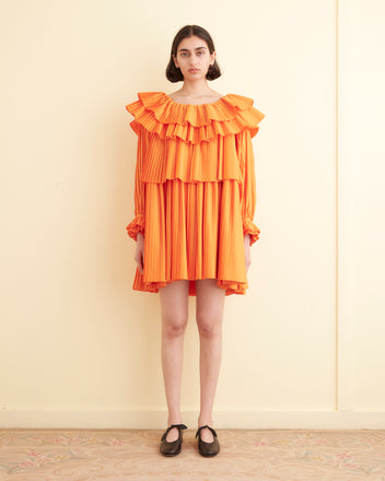 A person stands against a cream-colored wall, wearing the vibrant Brunch Dress - Orange. This 1970s-inspired dress features ruffled layers and is paired with black shoes. With shoulder-length dark hair, they look directly at the camera showcasing this chic and eco-friendly piece made from recycled polyester.