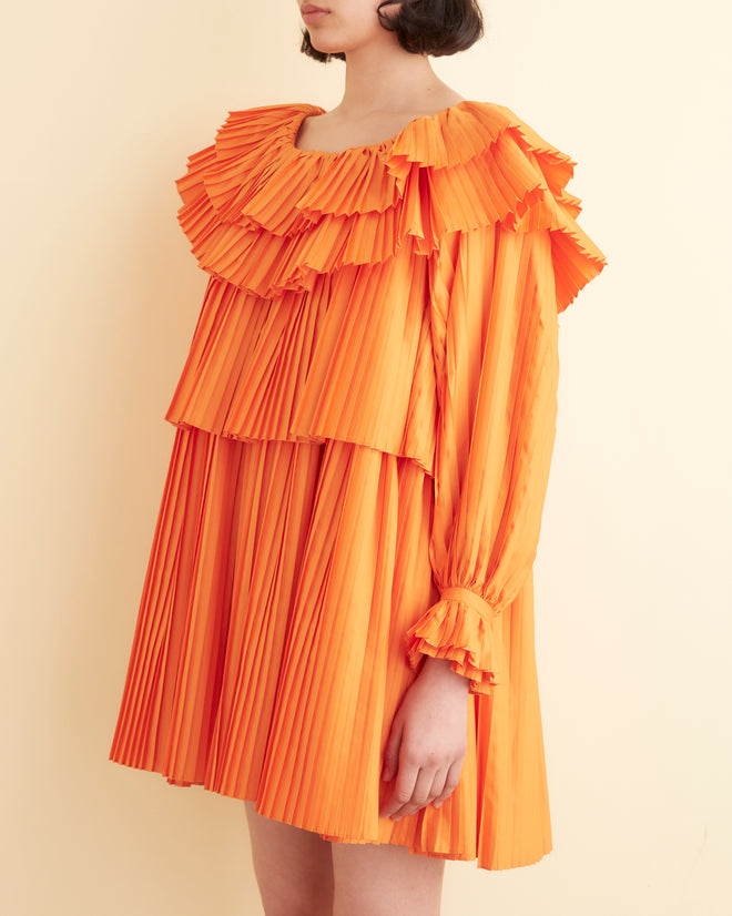 A person stands against a cream-colored wall, wearing the vibrant Brunch Dress - Orange. This 1970s-inspired dress features ruffled layers and is paired with black shoes. With shoulder-length dark hair, they look directly at the camera showcasing this chic and eco-friendly piece made from recycled polyester.