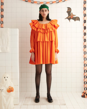 A person stands against a cream-colored wall, wearing the vibrant Brunch Dress - Orange. This 1970s-inspired dress features ruffled layers and is paired with black shoes. With shoulder-length dark hair, they look directly at the camera showcasing this chic and eco-friendly piece made from recycled polyester.