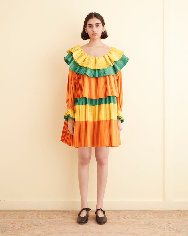 A person is standing in a room, wearing the Brunch Dress - Multi, a knee-length 1970s inspired dress featuring orange, yellow, and green ruffled tiers with long sleeves. They are also donning black shoes.