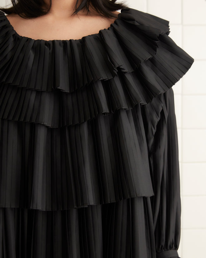 A person stands against a plain wall, wearing the Brunch Dress in black, which showcases pleated design and cascading layered ruffles paired with black flats. Made from recycled polyester, this elegant attire is perfect for any occasion. The setting appears indoors with a light-colored carpeted floor.