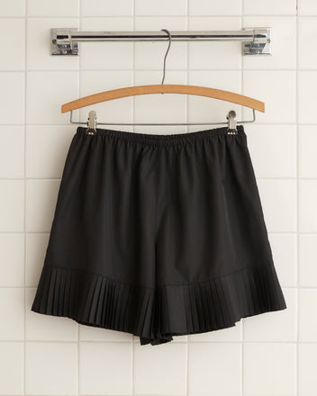 A pair of Brunch Shorts - Black, made from recycled polyester and featuring pleated hem detailing, hangs on a wooden hanger against a tiled wall, bringing a touch of 1970s fashion to your wardrobe.