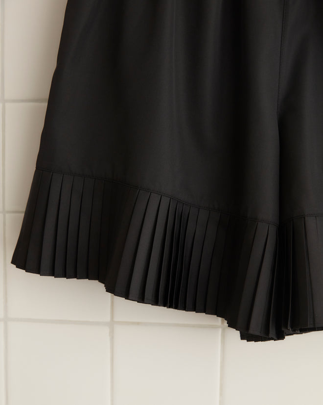 A pair of Brunch Shorts - Black, made from recycled polyester and featuring pleated hem detailing, hangs on a wooden hanger against a tiled wall, bringing a touch of 1970s fashion to your wardrobe.