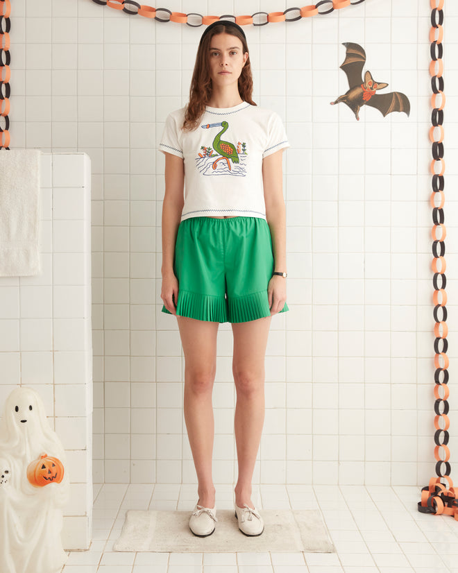 A woman stands in a tiled room, wearing a white graphic T-shirt, the Brunch Shorts in green made from 1970s-inspired recycled polyester, and white loafers. A folded towel is visible on the left.