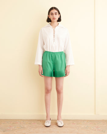 A woman stands in a tiled room, wearing a white graphic T-shirt, the Brunch Shorts in green made from 1970s-inspired recycled polyester, and white loafers. A folded towel is visible on the left.