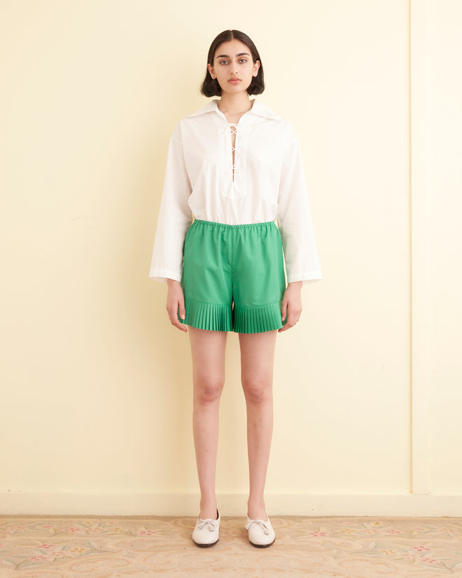 A woman stands in a tiled room, wearing a white graphic T-shirt, the Brunch Shorts in green made from 1970s-inspired recycled polyester, and white loafers. A folded towel is visible on the left.