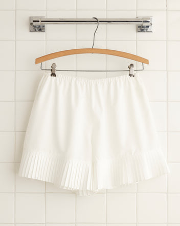 A pair of Brunch Shorts - White, crafted from recycled polyester, hangs on a wooden hanger against a white tiled wall.