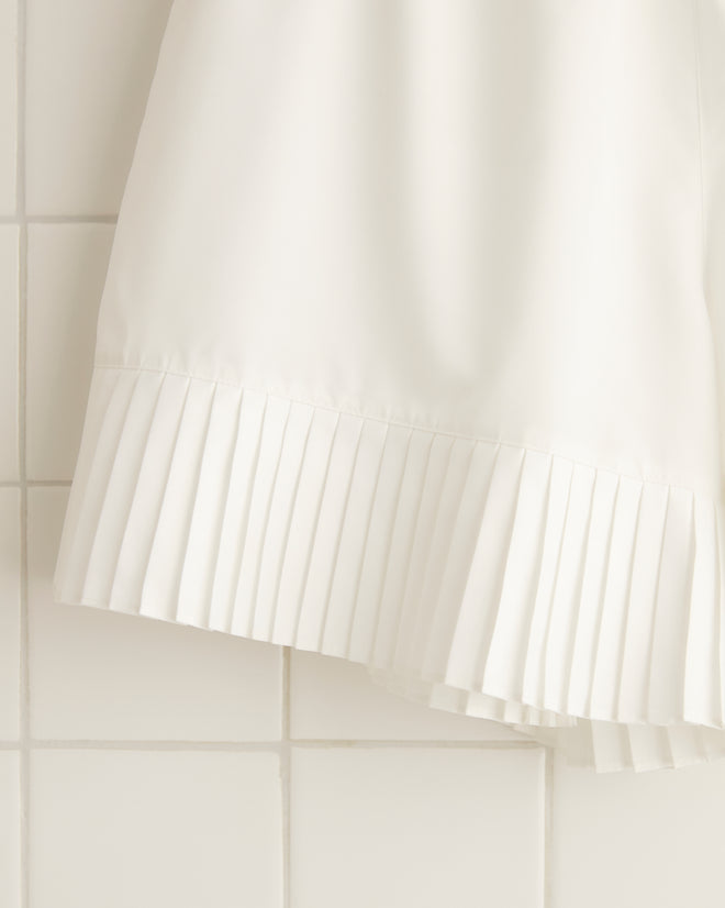 A pair of Brunch Shorts - White, crafted from recycled polyester, hangs on a wooden hanger against a white tiled wall.