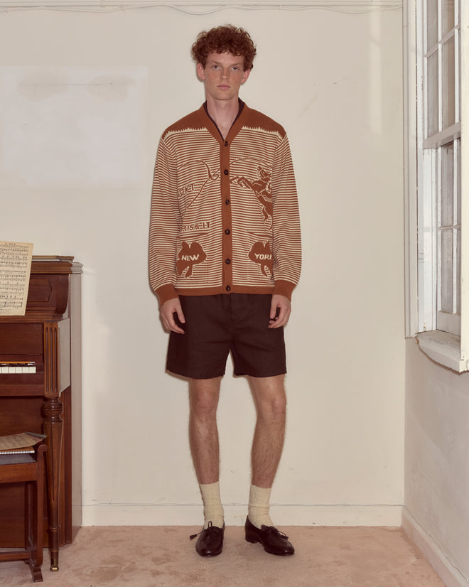 A man in a Buckaroo Cardigan and shorts stands indoors by a piano and a window.