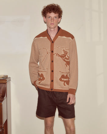 A young man with curly hair stands indoors by a piano, wearing the Buckaroo Cardigan and dark shorts.