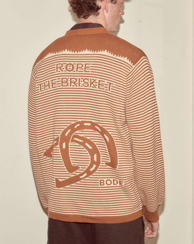 A man sports the Buckaroo Cardigan, a brown and beige striped cowboy sweater featuring "ROPE THE BRISKET" and a rope graphic on the back, inspired by team roping style.
