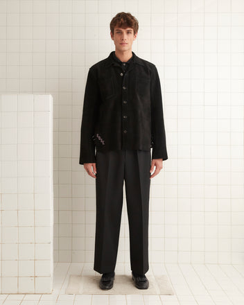 A man wearing a Buckled Suede Work Shirt in black, which features a subtle leather dickie, stands in a tiled room, facing forward with his hands by his sides.