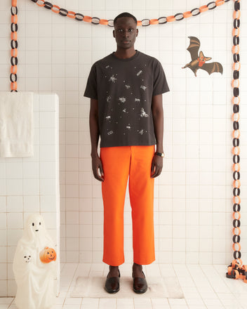 A person wearing the Bug Tee, a black Halloween-themed shirt made of 100% cotton with an insect print, and orange pants stands in a room decorated with paper chains and a ghost figure. The intriguing design of their shirt adds to the festive atmosphere.