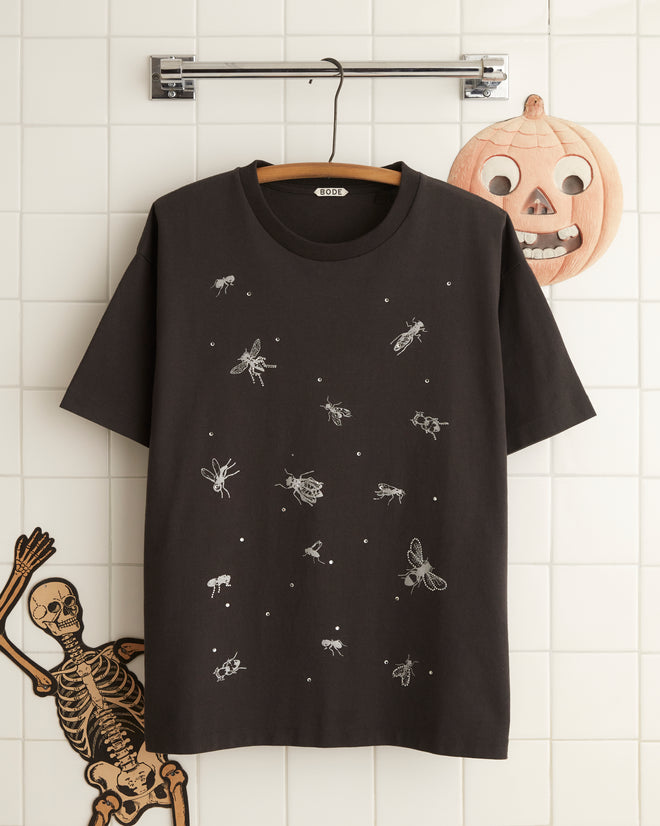 A person wearing the Bug Tee, a black Halloween-themed shirt made of 100% cotton with an insect print, and orange pants stands in a room decorated with paper chains and a ghost figure. The intriguing design of their shirt adds to the festive atmosphere.