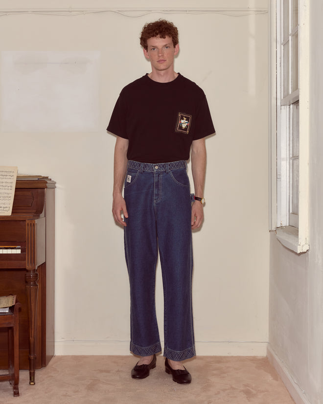 A man in a black Bullion Card Tee and wide-leg jeans stands indoors by a piano and window, looking at the camera.
