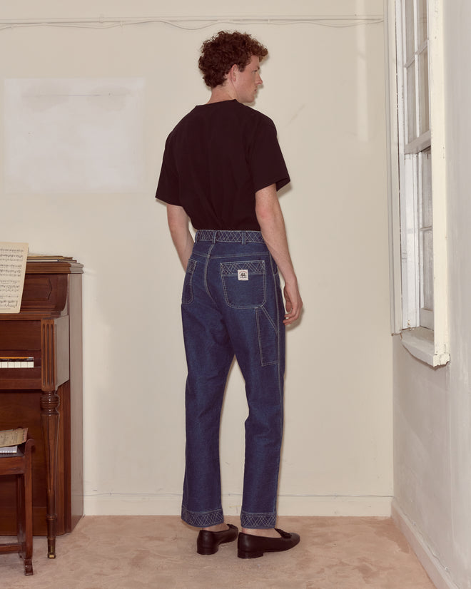 A person in a Bullion Card Tee and blue jeans stands facing a corner near a piano and window in a beige room.