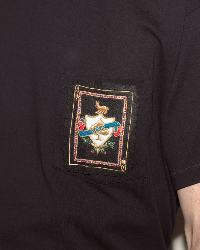 Close-up of the Bullion Card Tee, a black 100% cotton shirt featuring an ornate embroidered patch with a shield and blue banner on the pocket.