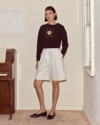 A woman stands indoors by a piano, looking at the camera while wearing a Bullion Card Pullover and white shorts.