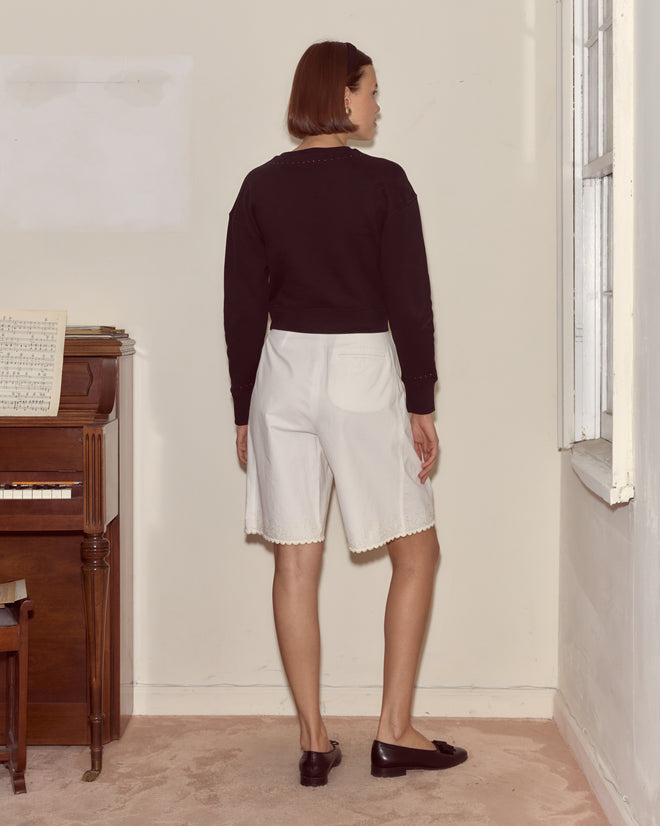 A woman stands indoors, facing away near a piano and window, wearing the Bullion Card Pullover and white shorts—a scene reminiscent of Alice’s Adventures in Wonderland.