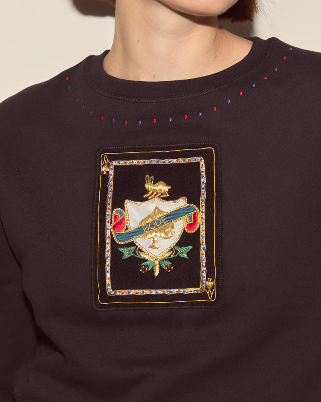 A person wears the Bullion Card Pullover, a dark shrunken top featuring a colorful embroidered crest, gold accents, and bullion appliqué on the chest.