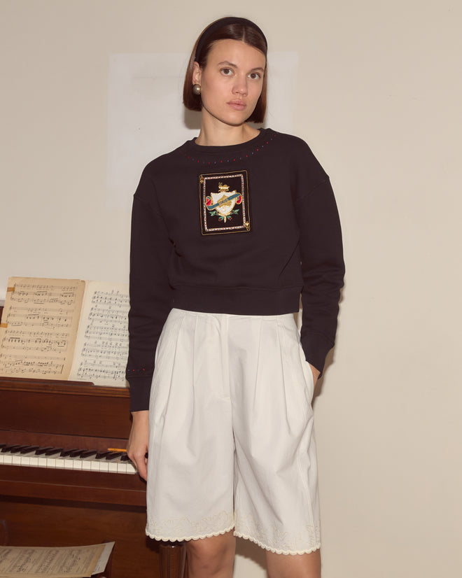 Woman wearing the Bullion Card Pullover with delicate bullion appliqué stands by a piano with sheet music, looking at the camera.