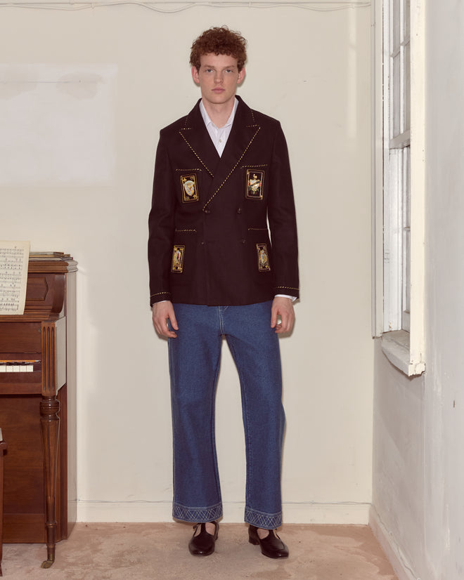 A man stands indoors wearing the Bullion Card Suit Jacket—a black blazer with metallic striped piping and gold details—paired with blue wide-leg jeans and black shoes.