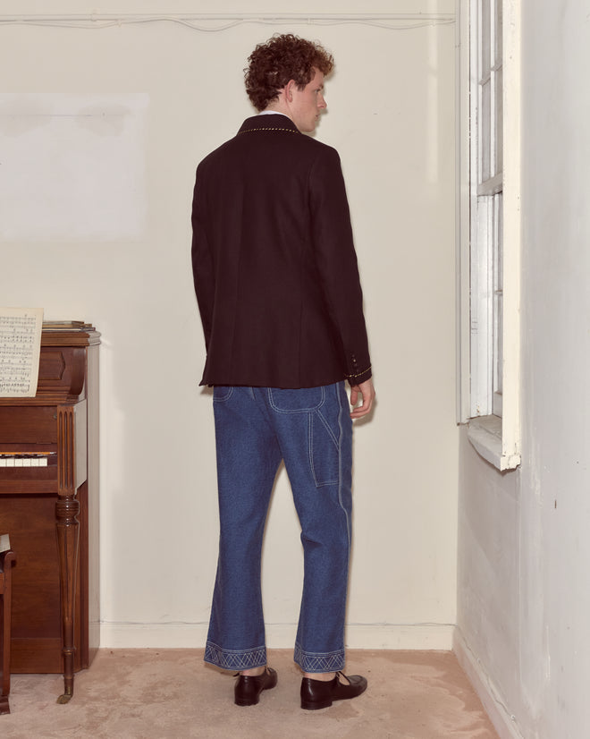 A person in the Bullion Card Suit Jacket with metallic striped piping and blue jeans stands facing a window in a room with a piano.