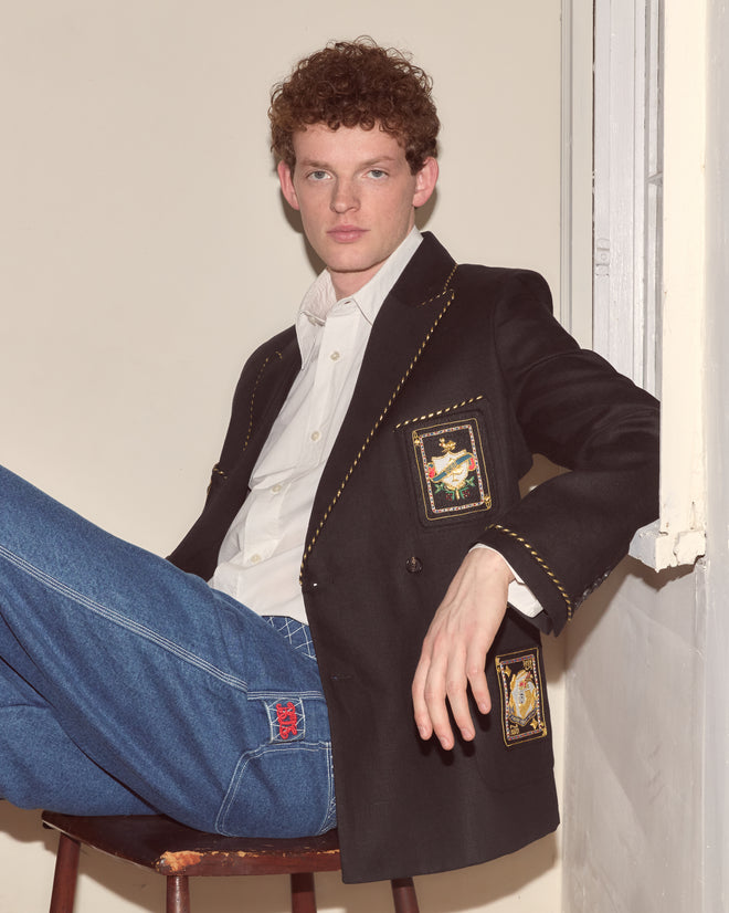 A person with curly hair wearing the Bullion Card Suit Jacket and jeans sits on a stool by a window, looking at the camera.