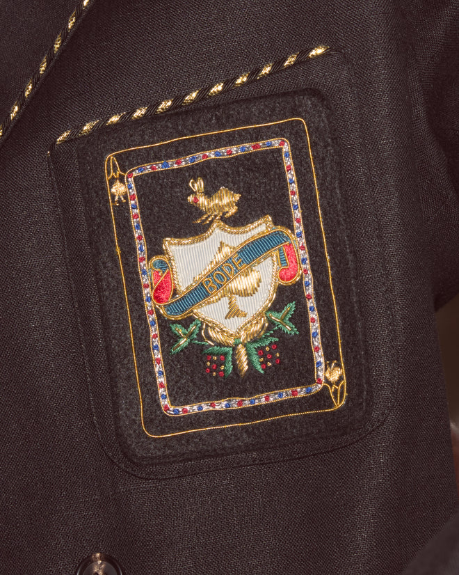 The Bullion Card Suit Jacket features a black blazer pocket with metallic striped piping and an ornate embroidered crest that includes a shield and a ribbon labeled "ADDE.