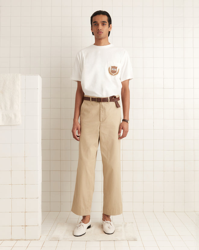 A person in a Sportsman Patch Pocket Tee and beige pants stands against a tiled wall, exuding an air reminiscent of early 20th-century style.
