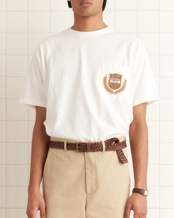 A person in a Sportsman Patch Pocket Tee and beige pants, with a braided brown belt, stands against a tiled wall.