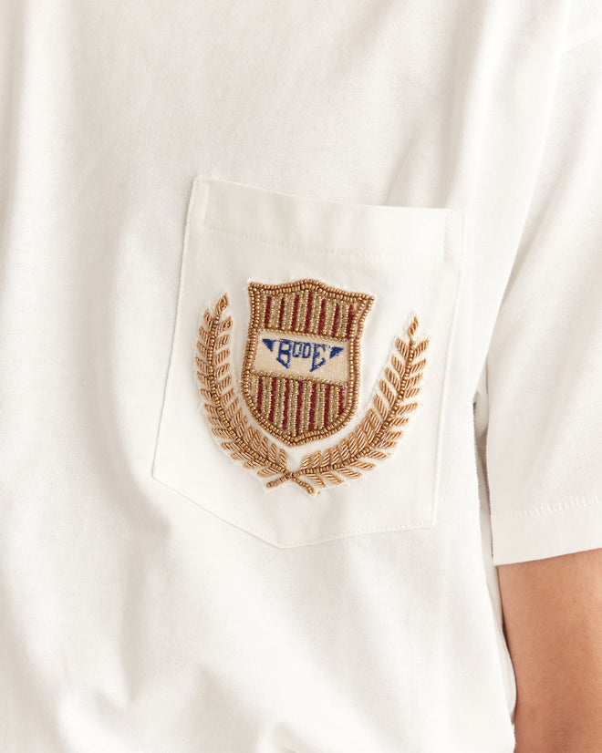 The Sportsman Patch Pocket Tee is a 100% cotton white shirt, featuring an embroidered shield and "Bode" text above a chest pocket, elegantly accentuated by gold laurel branches reminiscent of early 20th-century designs.