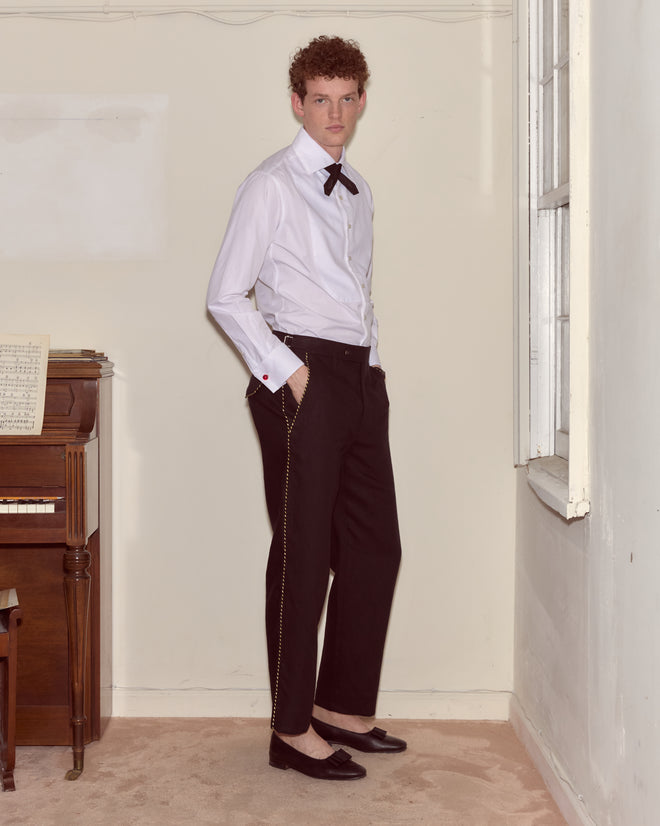 A man in a white shirt, Bullion Piping Trousers, and loafers stands indoors beside a piano and a window.