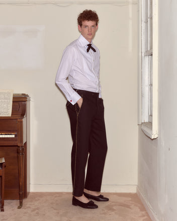 A man in a white shirt, Bullion Piping Trousers, and loafers stands indoors beside a piano and a window.