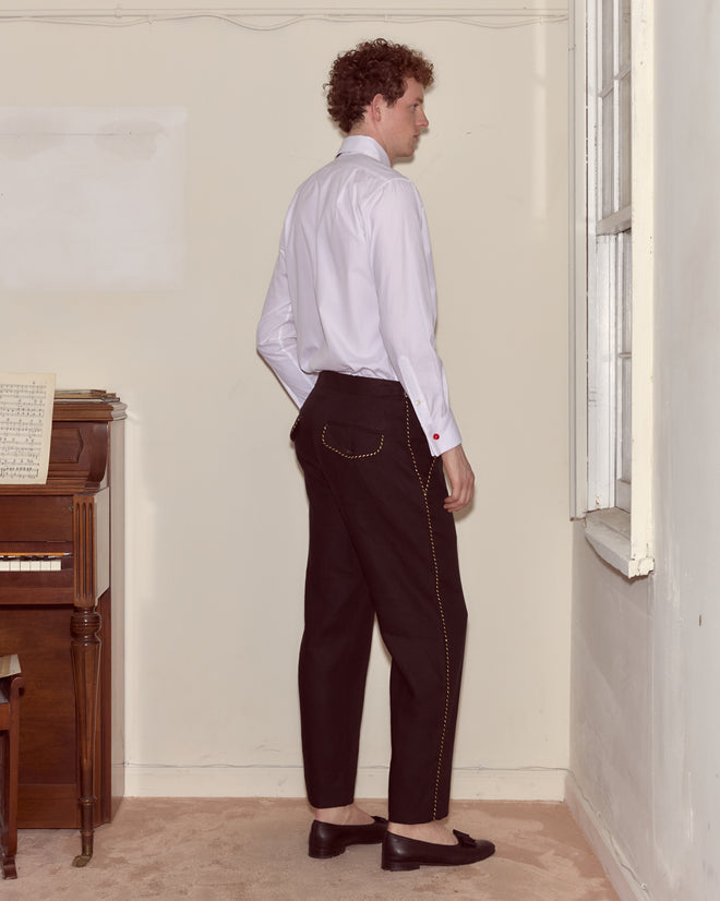 A person in a white shirt and Bullion Piping Trousers stands sideways near a piano in a sparsely furnished room.