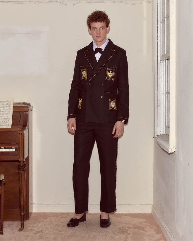 A man in Bullion Piping Trousers, styled with gold details and striped metallic piping, stands by a piano and window in a warmly lit room.