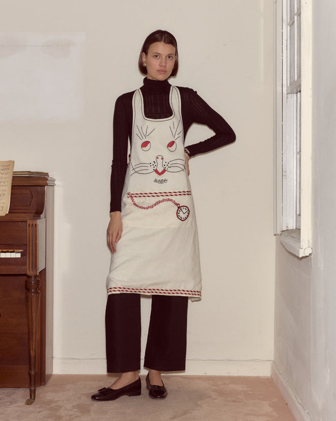 A woman in black clothing wears the Bunny Apron, featuring a bunny face design, while standing by a piano in a neutral room.