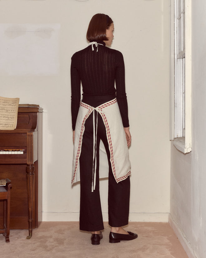 A woman in black clothes and loafers faces a wall, wearing the Bunny Apron, a white apron tied at the back.