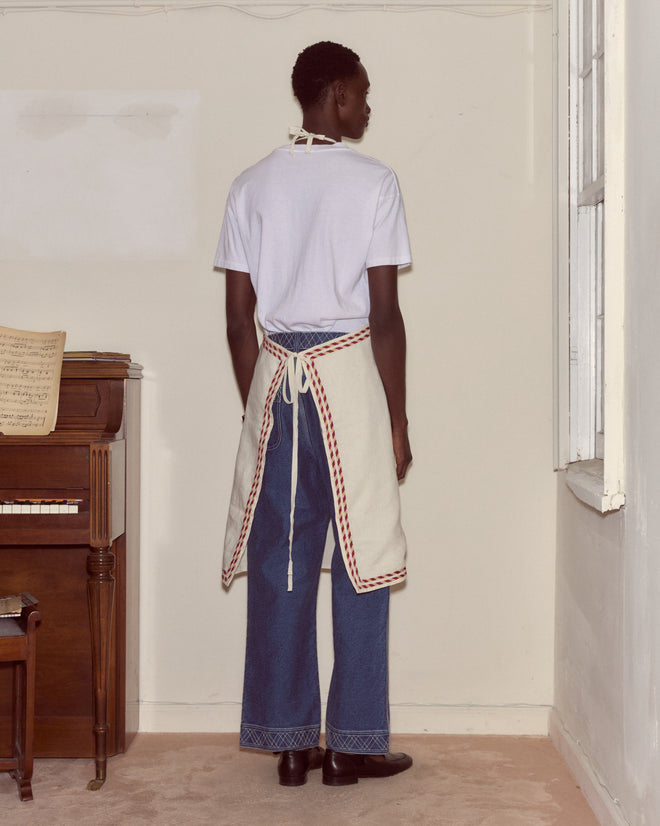 A person in a white shirt, jeans, and a Bunny Apron stands facing a wall near a piano and a window.