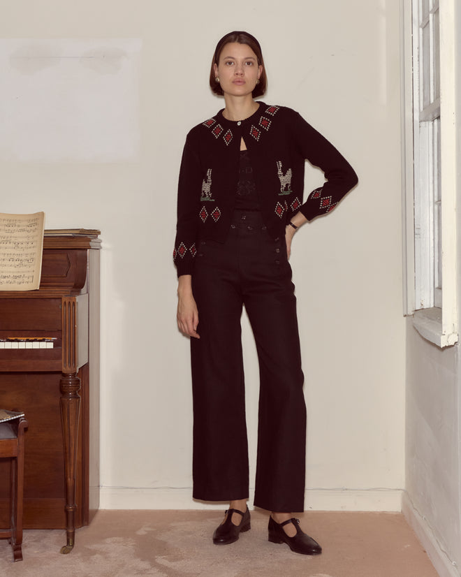 A woman in a Bunny Cardigan and black pants stands by a piano and window in a softly lit room, her 100% wool knitwear with rabbit designs adding whimsy to the cozy setting.