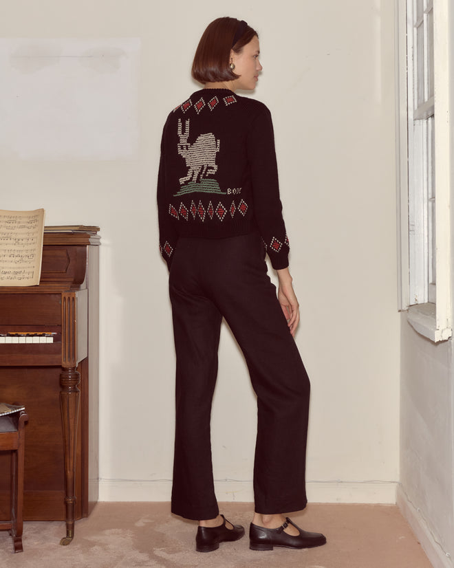 Woman in a black Bunny Cardigan featuring a white bunny design, standing by a piano and looking out a window.